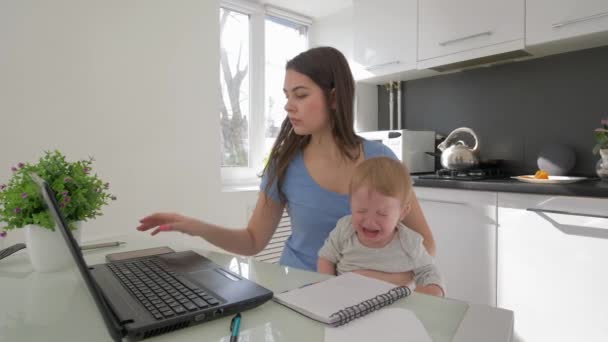 Combinando el trabajo y la crianza, la madre con el niño llorando mientras trabaja en el ordenador portátil y hablando en el teléfono móvil sentado en la mesa en la cocina — Vídeos de Stock