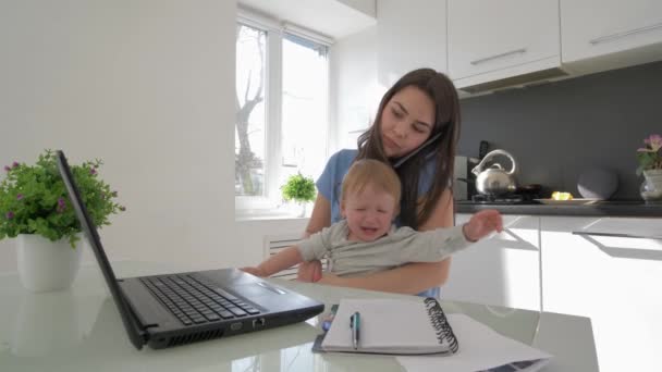Family stress, multitasking mum with crying infant boy while working on laptop computer and talking on cell phone sitting at table in kitchen — Stock Video