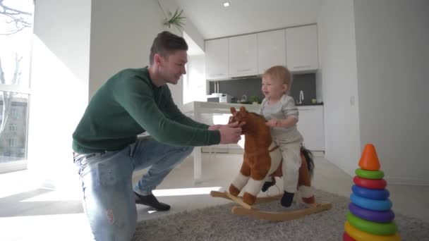 Juegos con los niños, padre feliz jugando con el hijo lindo en asientos de caballo de peluche y balanceo en casa en la cocina — Vídeo de stock