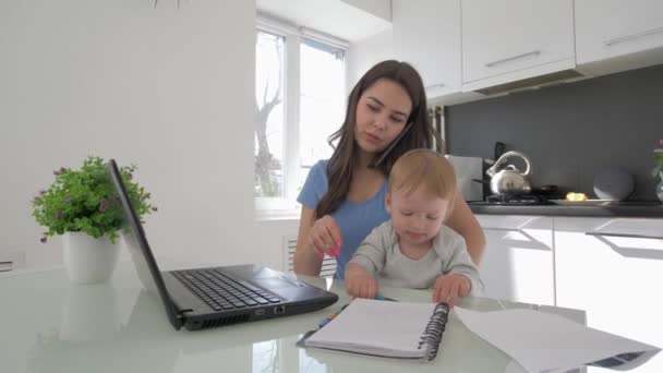 La paternidad dura, mamá multitarea con niño llorando niño mientras trabaja en el ordenador portátil y hablando por teléfono sentado en la mesa en la cocina — Vídeo de stock