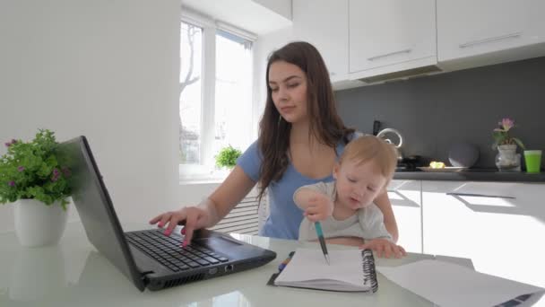 Wielozadaniowe macierzyństwo, biznes kobieta połączyć wychowanie chłopca i pracy na laptopie komputera i rozmowy na telefon komórkowy siedzi przy stole w kuchni — Wideo stockowe