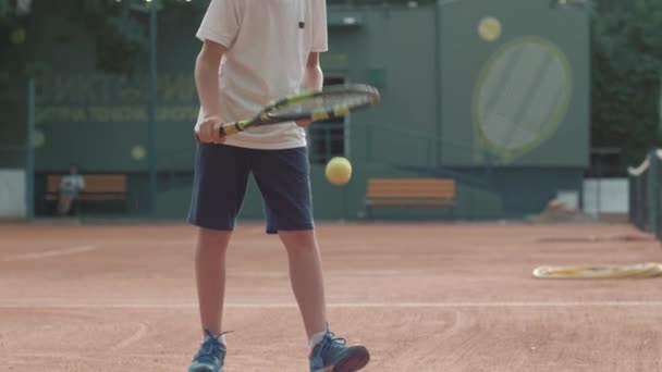 Portrait of boy tennis player with racket while stuffing ball on ground on tennis court on open air — ストック動画