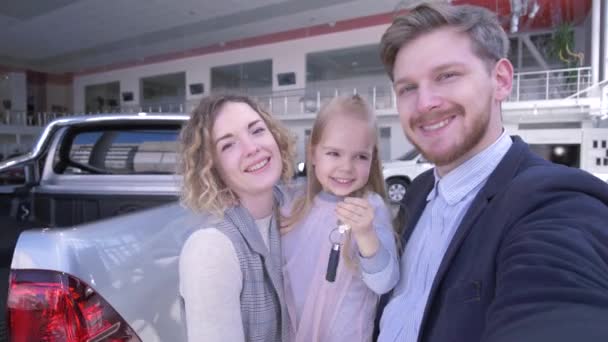 Photo selfi de famille dans le centre automobile, les parents avec fille enfant mignon avec des clés prennent selfie photo sur téléphone portable près de nouveau véhicule acheté chez le concessionnaire automobile — Video