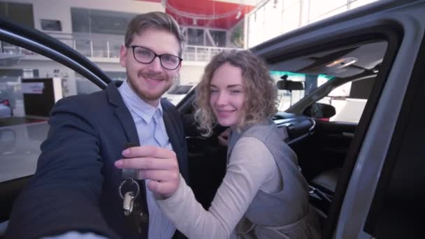 Bloggers guy with girl with keys record video in live broadcast on mobile phone near new purchased automobile in car showroom — Stock Video