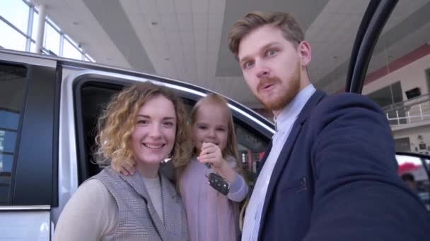 Foto selfi familia en la tienda de automóviles, pareja divertida con linda niña con llaves fotografiadas en el teléfono inteligente cerca del nuevo automóvil comprado en el centro de venta de coches — Vídeo de stock