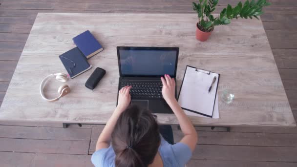 Γενικά άποψη της freelancer κορίτσι πληκτρολογώντας κείμενο στο πληκτρολόγιο laptop και γράφει σημειώσεις στο πρόχειρο κάθεται στο τραπέζι κατά τη διάρκεια της μελέτης σε απευθείας σύνδεση — Αρχείο Βίντεο
