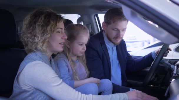 Araba işi, mutlu gülümseyen anne ve baba ile küçük kız aile otomobili gözden geçiriyorlar. Araba galerisinde kamarada otururken el sallıyorlar ve başparmak sallıyorlar. — Stok video