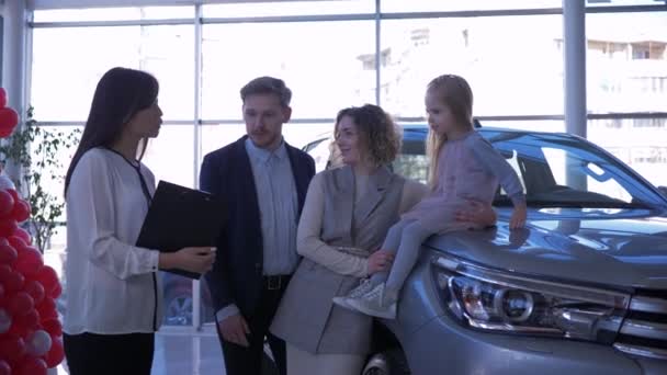 Автосалон, молодые покупатели семьи с ребенком девушка общаться с автомобильным менеджером о покупке нового автомобиля в центре автопродажи — стоковое видео