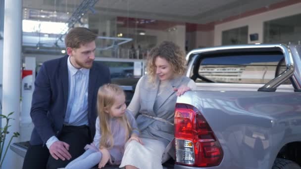 Centro de venta de automóviles, pareja joven con niño elige automóvil y se comunican entre sí mientras están sentados en el maletero en la sala de exposición del coche — Vídeo de stock
