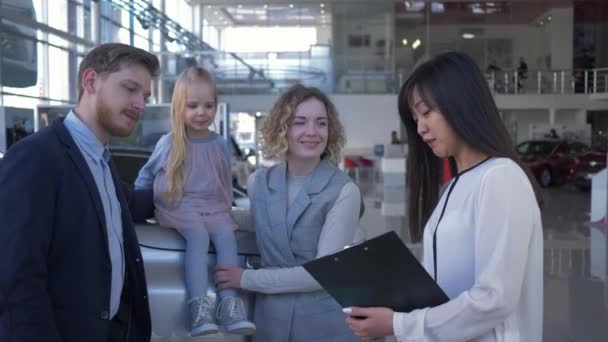 Auto vendeuse conseille les clients famille avec enfant sur le capot à l'achat de voiture dans le showroom — Video