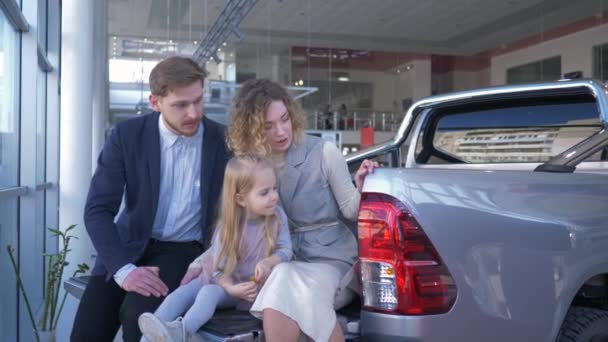 Tienda de automóviles, retrato de la familia feliz con la hija elige el coche y consultar entre sí mientras está sentado en el maletero en el centro de ventas — Vídeos de Stock