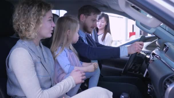 Família alegre com a menina pequena da criança está feliz em comprar o carro enquanto examina o automóvel no salão no centro de auto — Vídeo de Stock