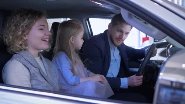 Família escolhe carro novo, mãe feliz e pai com criança menina considerando automóvel sentado na cabine no salão de auto — Vídeo de Stock