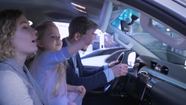 Семья в автосалоне, счастливые родители с маленькой девочкой душат машину и сдают большие пальцы, сидя в салоне в автосалоне — стоковое видео