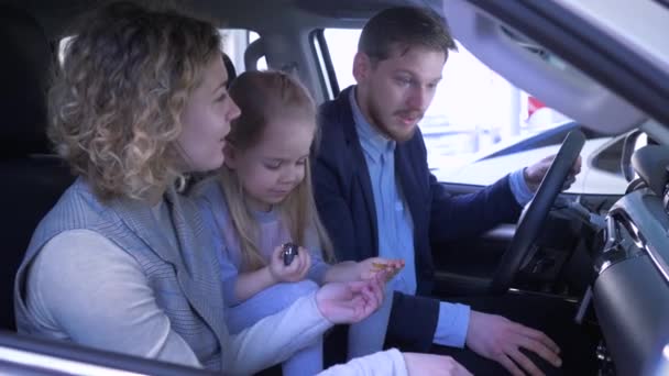 Familj i bil försäljningscentrum, glad leende mamma och pappa med unge flicka recensioner bil och vifta nyckel när du sitter i kabinen på auto återförsäljare — Stockvideo