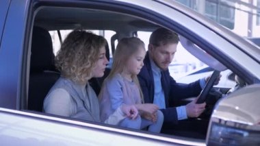 Otomobil satış merkezindeki mutlu müşteri, çocuklu gülümseyen aile yeni arabayı denetliyor ve oto tamirhanesinde kabinde otururken olumlu hareket ediyor.