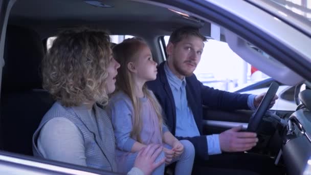Zadowoleni klienci w warsztacie samochodowym, uśmiechnięta rodzina z córeczką wybiera samochód rodzinny machając kluczykiem i dając pozytywny gest siedząc w kabinie w salonie samochodowym — Wideo stockowe