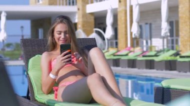 Yaz tatili, elinde cep telefonu olan neşeli ve çekici bir kız tatil sırasında pahalı otellerde havuz başında dinleniyor.