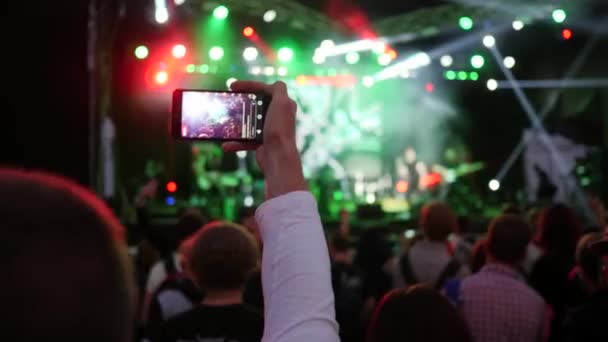 Вентилятор со смартфоном в руке записывает видео рок-фестиваля в ночное время в ярких огнях сцены — стоковое видео