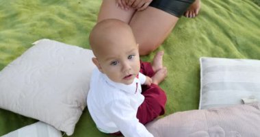 Bebek takım elbiseli ve kocaman gözleri var. Dışarıdaki parktaki yastıkların yanında yeşil ekose üzerinde oturan kameraya bakıyor.