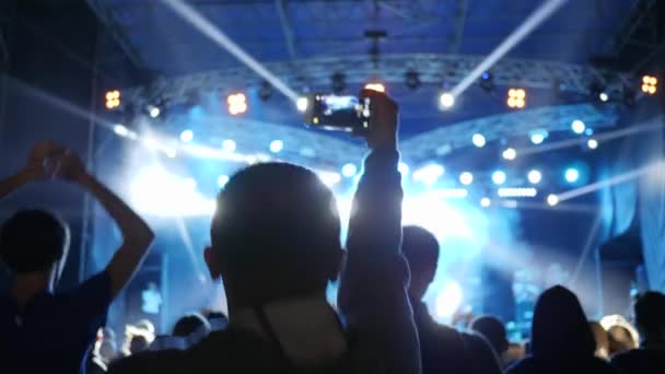 Homem pulando com smartphone em mão grava vídeo no fundo multidão em luzes de palco brilhantes no concerto — Vídeo de Stock