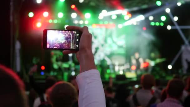 Фан з смартфоном в руці записує відео на фоні натовпу в яскравих фарах рок-фестивалю — стокове відео