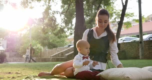 Joven madre se sienta con lindo bebé en traje y pajarita en el césped verde en el parque — Vídeos de Stock