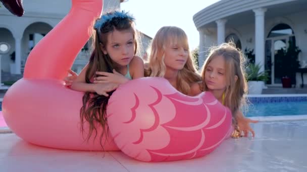 Niñas se acuestan en flamenco rosa inflable cerca de la piscina, celebridades de los niños en traje de baño en las vacaciones de verano, niños ricos estropeados — Vídeos de Stock