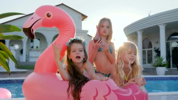 Las muchachas dulces mienten en el flamenco rosado inflable cerca de la piscina, niños ricos estropeados en retroiluminación al aire libre, niños se divierten — Vídeos de Stock