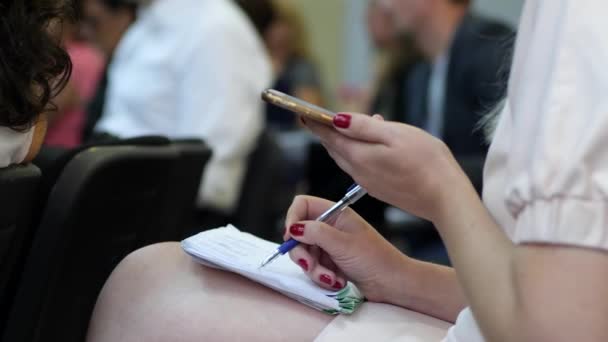 Mão feminina escreve algo no bloco de notas usando telefone celular na sala de reuniões — Vídeo de Stock