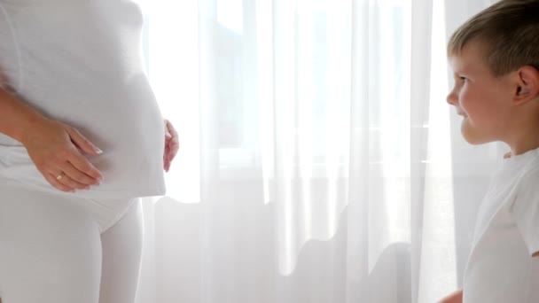 Любопытный ребенок прикасается и слушает живот беременной женщины в белой одежде — стоковое видео