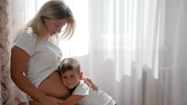 Little son hugs expectant female and smiling on background of white curtains — Stock Video