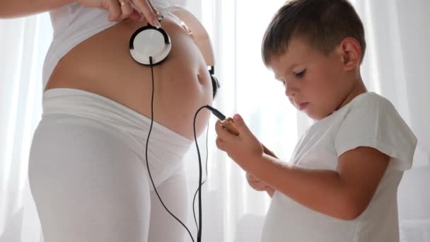 Niño pequeño con teléfono móvil en la mano poner auriculares en el vientre de la mujer embarazada — Vídeos de Stock