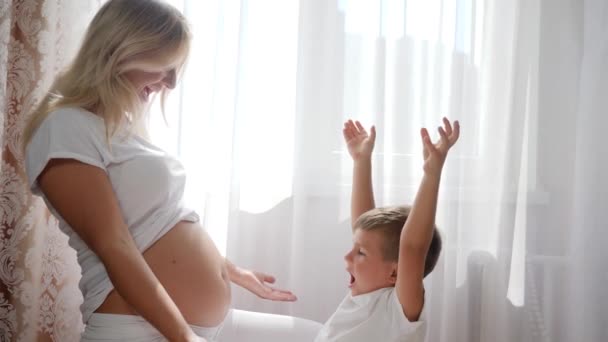 Kind legt Kopf auf großen Bauch der Frau, die Baby im Zimmer erwartet, auf Hintergrund weißer Vorhänge — Stockvideo