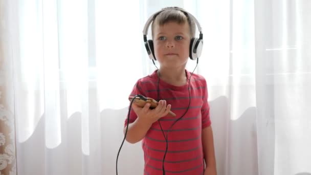 Enfant souriant avec téléphone dans les mains en écoutant de la musique à travers le casque et en dansant sur fond de rideaux blancs — Video