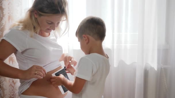 Jövendőbeli anya és kisfia a leendő gyermek szkennelésével a kezében fehér függönyök hátterében — Stock videók