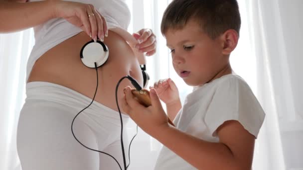 Niño pequeño con el teléfono móvil en la mano poner los auriculares en el vientre de la mujer embarazada en la habitación iluminada — Vídeos de Stock