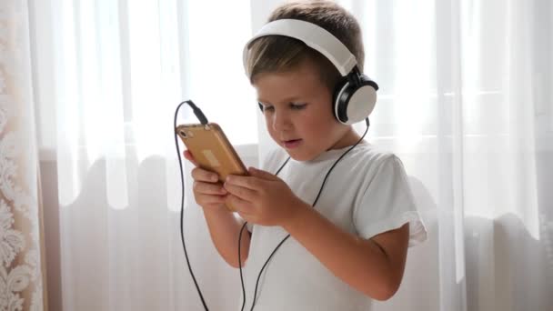 Glimlachend kind met mobiel in de arm genieten van muziek in koptelefoon en dansen in de kamer — Stockvideo