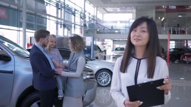 笑顔の車のディーラーの肖像自動車販売センターで自動車を購入した所有者の子供と幸せな若いペアを背景にアジアの女性 — ストック動画
