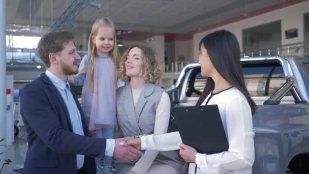 成功した車の販売、幸せな女性マネージャーは自動車ショールームで自動車の家族のバイヤーと握手し、カメラを見て笑っています — ストック動画