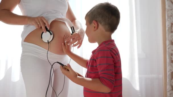 Mujer en ropa blanca da música al bebé en el vientre a través de auriculares, teléfono móvil en manos de niño pequeño — Vídeos de Stock