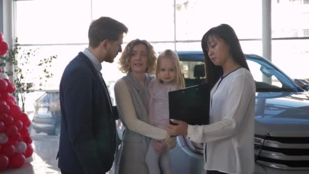 Pembelian kendaraan, pasangan bahagia dengan anak kecil menginformasikan dengan konsultan mobil tentang pembelian mobil keluarga di showroom penjualan — Stok Video