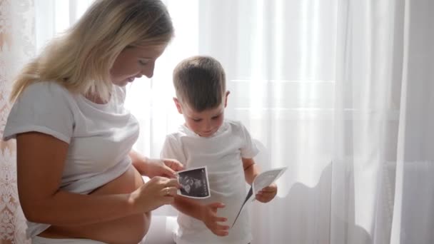 Молода вагітна мати з милою дитиною дивиться фото-сонографію вдома в яскравому природному світлі — стокове відео