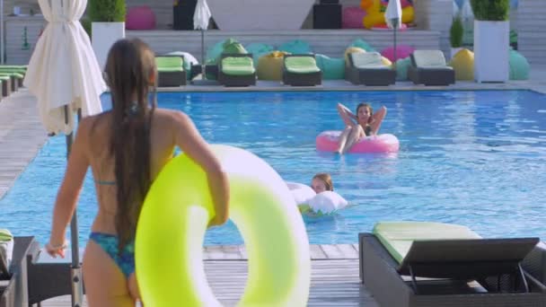 Amigos felices en la piscina de verano, las mujeres jóvenes se relaja en la piscina y la novia con anillos inflables salta al lado del agua con aerosol durante las vacaciones en el complejo — Vídeos de Stock