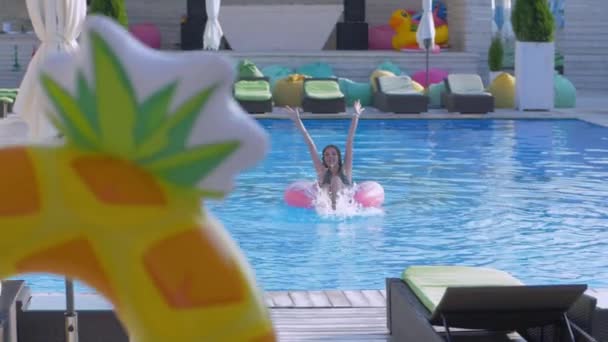 Descanso de verano, la joven feliz se relaja en anillos inflables en la piscina y la novia salta al lado del agua con aerosol durante el fin de semana en el resort — Vídeo de stock