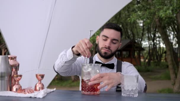 Kocsmáros a bárpulton keveri az alkoholt jéggel az üvegben, csapos csinál alkoholos italt a szabadban, nyári kávézó — Stock videók
