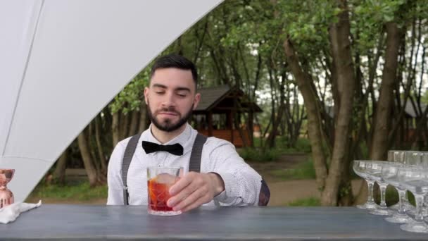 Бармен показує жест схвалення, великий палець бармена, усміхнений бармен подає віскі з льодом на барній стійці, літній ресторан — стокове відео