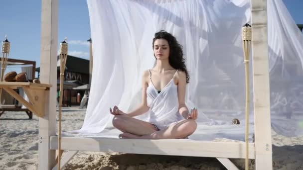 Hembra en posición de loto meditación en bungalow, yoga en la playa en verano, viento desarrolla pelo y tela blanca, islas tropicales — Vídeo de stock