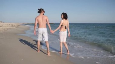 Romantik yürüyüş yapan pozitif genç insanlar, kumların üzerinde çıplak ayakla yürüyen arkadaşlar, aşık çiftler egzotik bir tatil, rüzgar kadınların saçlarını büyütüyor.