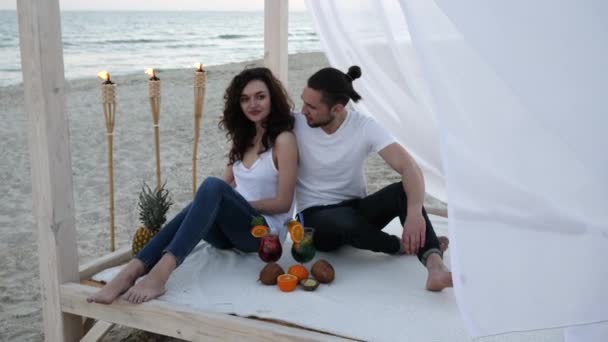 Feliz casal com coquetéis, divirta-se, vento desenvolve pano branco, bungalow, umas férias exóticas, no fundo frutas tropicais — Vídeo de Stock